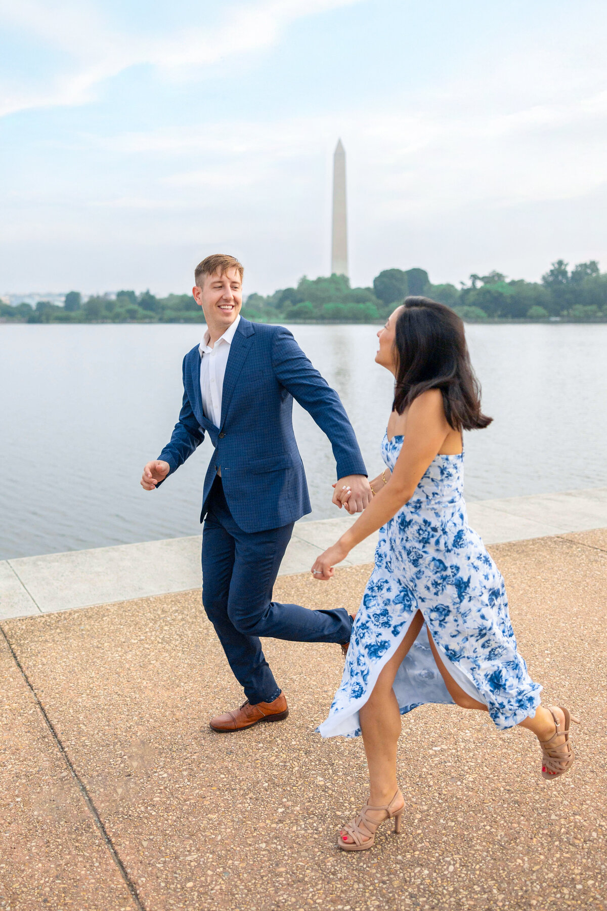 A couple holding hands, smiling, and running along a pathway by a body of water. The man is in a blue suit, and the woman is wearing a blue and white floral dress. The Washington Monument is visible in the background.
