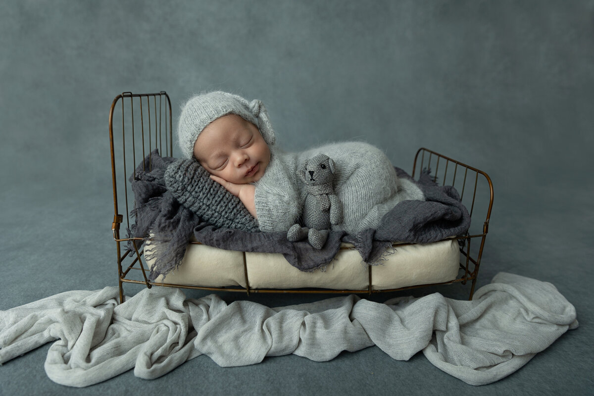A newborn baby sleeps in a metal bed with a stuffed bear in a NJ Newborn Photography studio