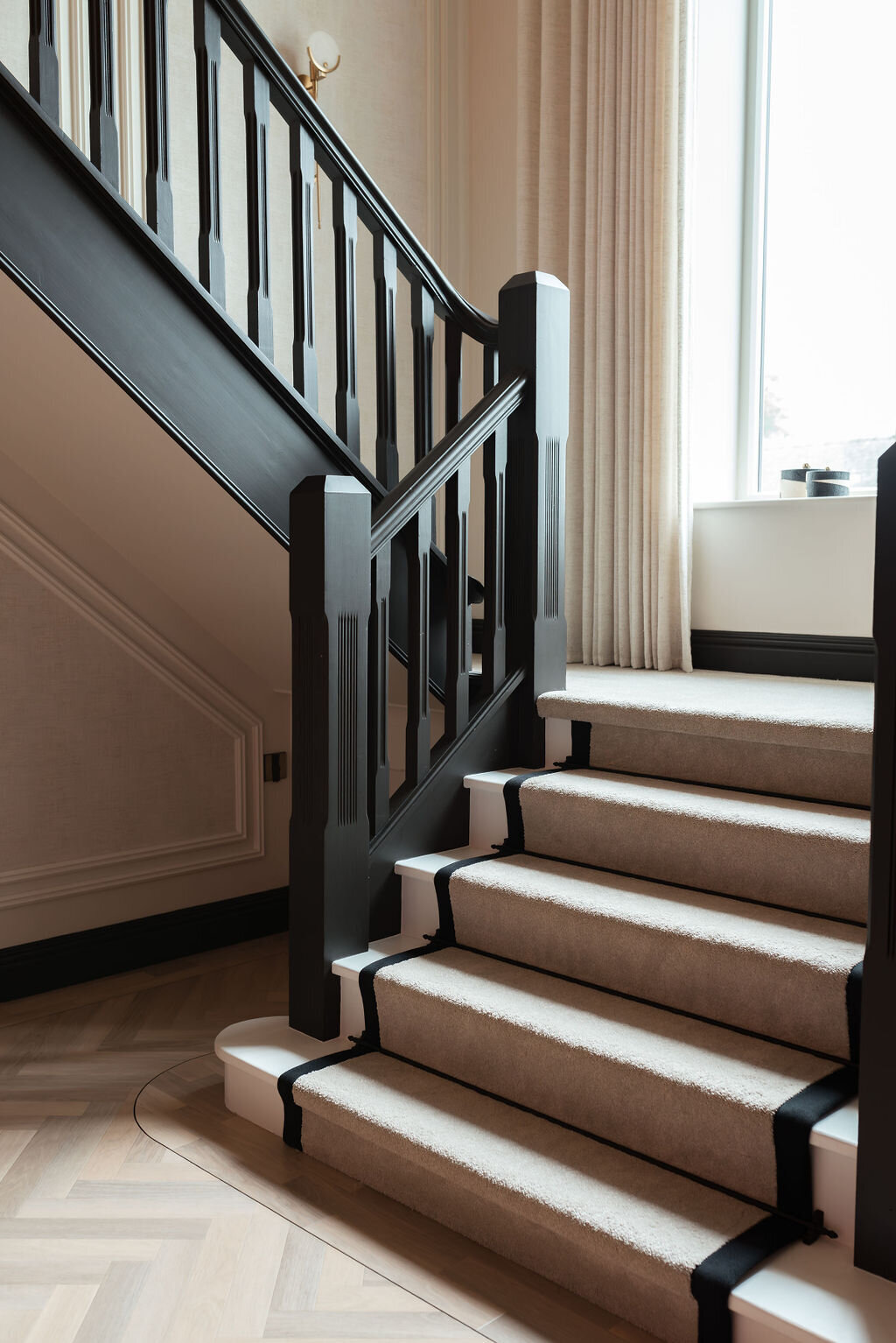 A modern, well-lit white staircase with black railings and beige carpet stair runner with black stripes leads up to a large window. Light-colored curtains frame the window.