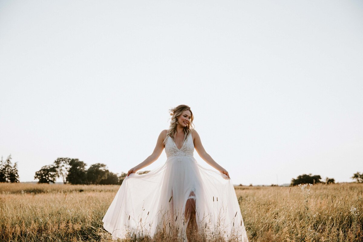 Bride standing in Exeter, Ontario wheat field for rustic farm wedding. She is holding her organza skirt with the light filtering through it.