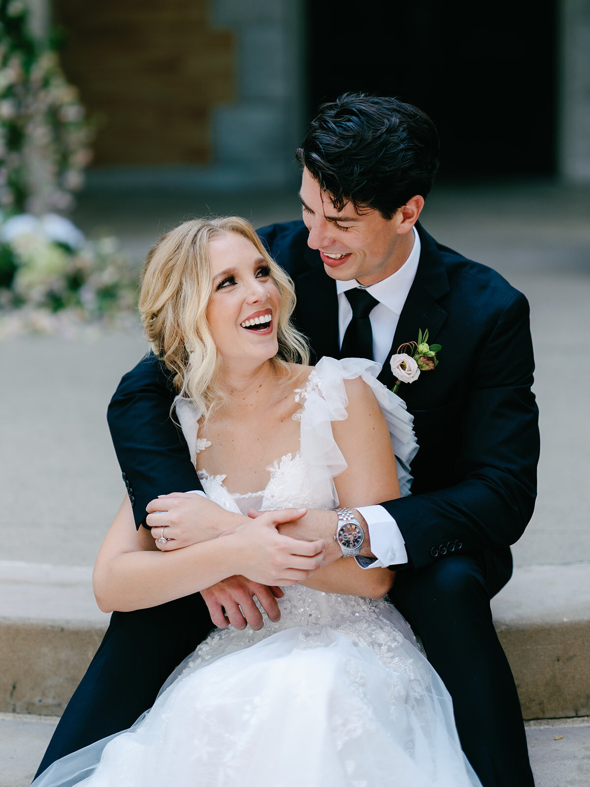 Bride and groom look at each other as they sit on the steps and they laugh at each other with joy.