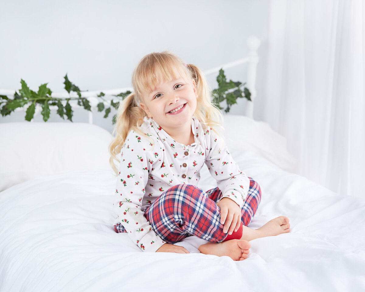 Image of a girl on a white bed with holly around the headboard with Christmas backdrop in the studio. Taken by Norwich portrait photographer Claire Howes. Christmas Mini Sessions Norwich.