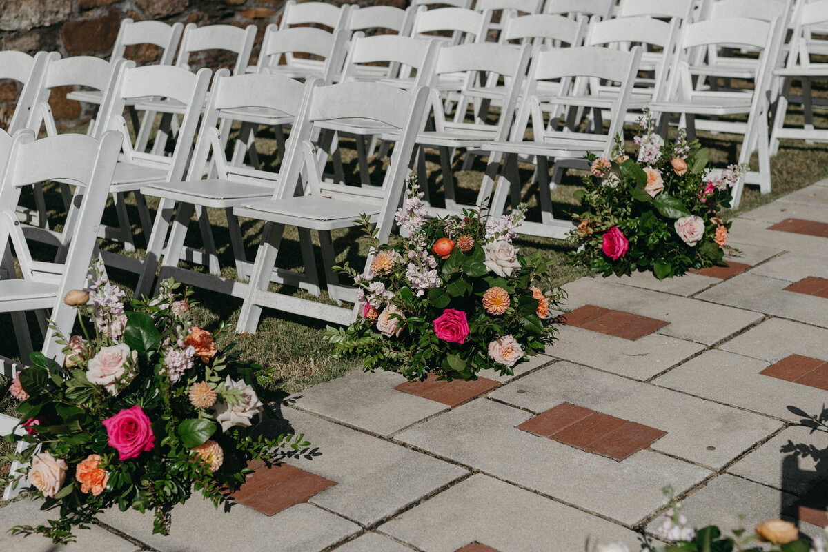 Ground meadow floral arrangements of roses, dahlias, and ranunculus lining the wedding ceremony aisle to create a colorful garden-inspired entrance.

