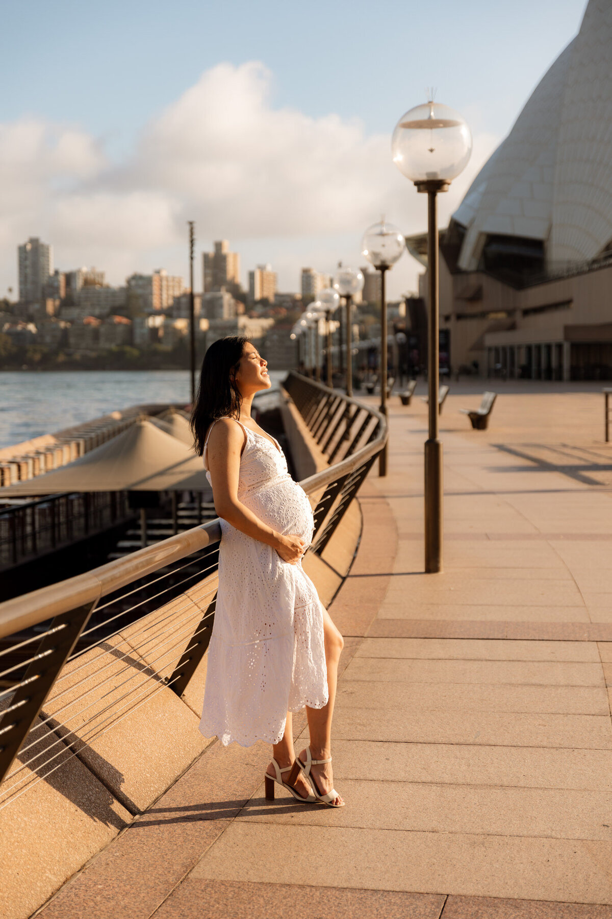 Pregnant woman in white dress enjoying golden hour sunlight near the Sydney Opera House during a maternity photoshoot