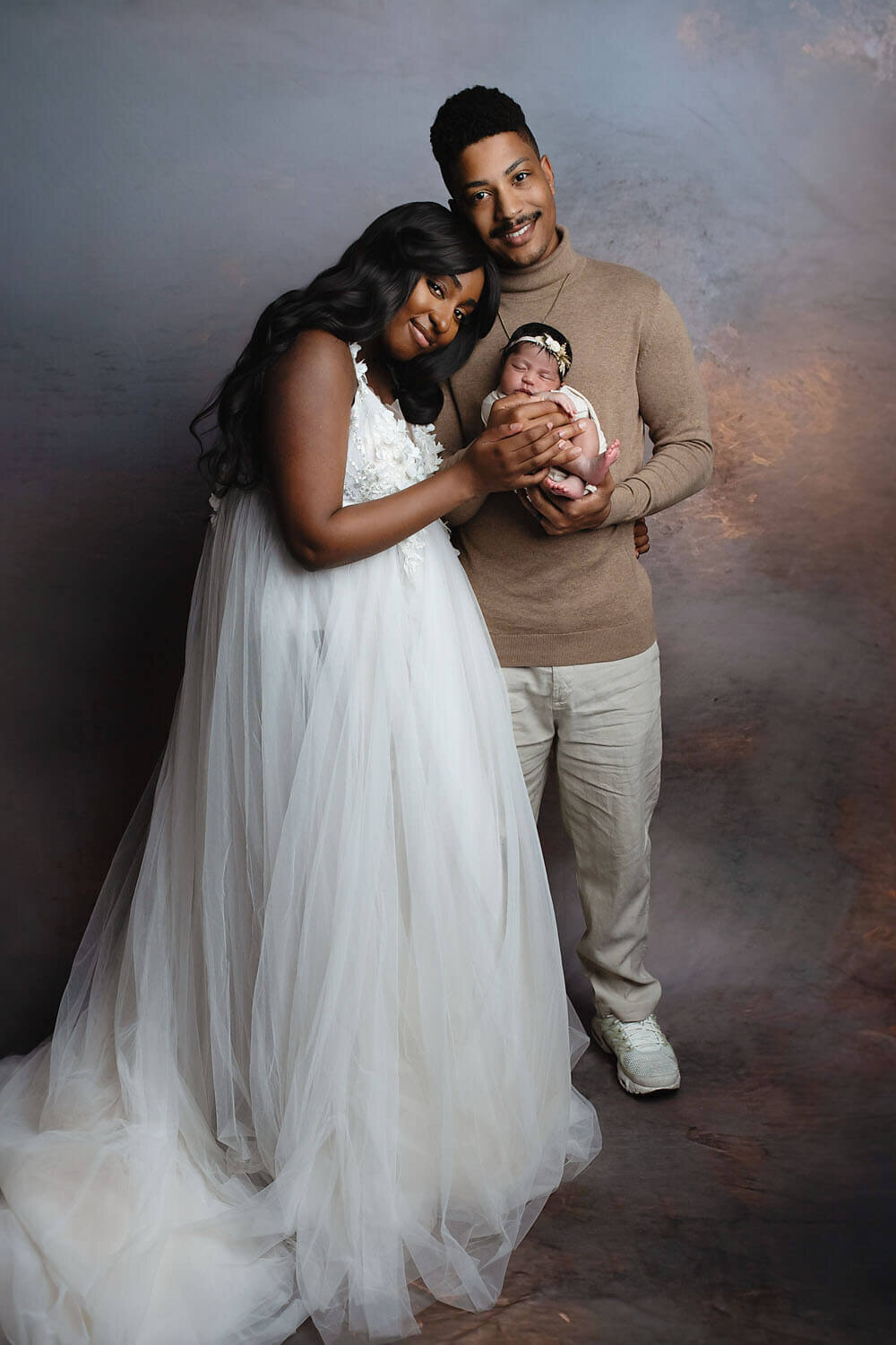 Happy parents stand in a studio smiling while dad cradles their sleeping newborn baby