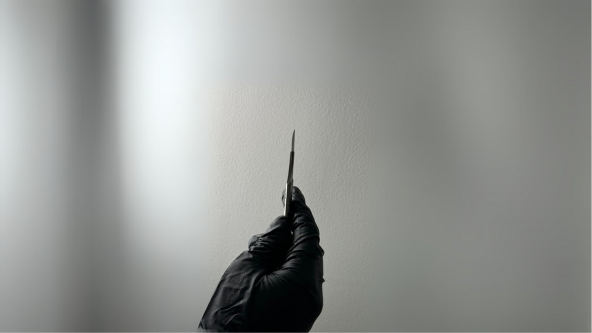 Image shows a hand  in a clinical black glove holding a dermaplaning scalpel against a white wall