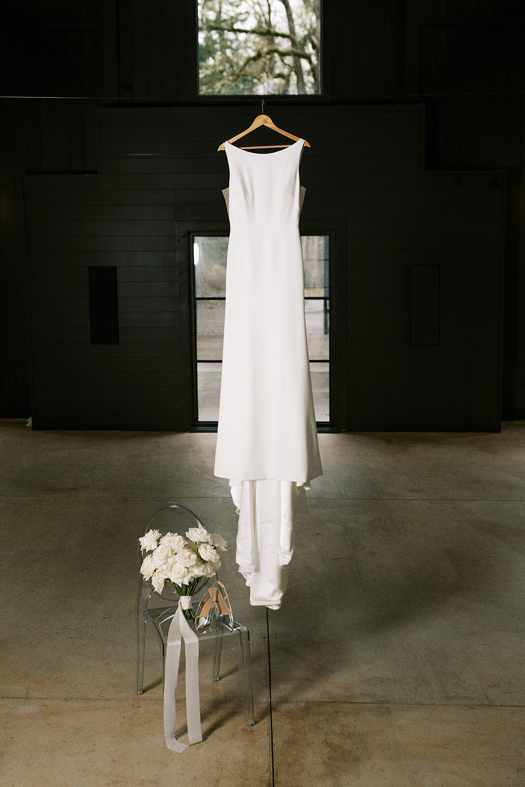 A wedding gown hanging next to a chair with a bouquet and shoes.