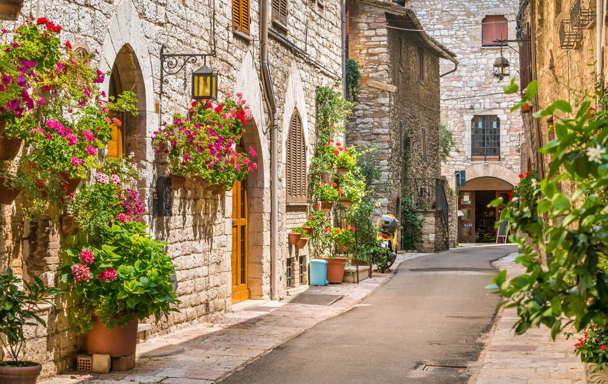 A charming narrow street in a historic town with stone buildings adorned with vibrant potted flowers. Flowering plants hang on walls, and colorful foliage lines the pathway, creating a picturesque, inviting atmosphere.