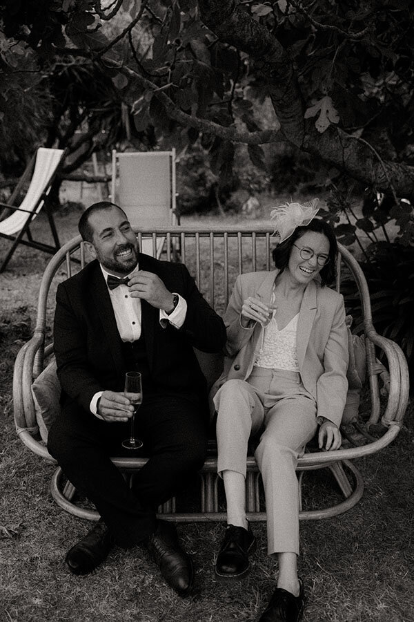Un couple d'invités rit, assis dans le jardin. Un moment de détente capturé en noir et blanc par Kamescop, vidéo et photographie de mariage.
