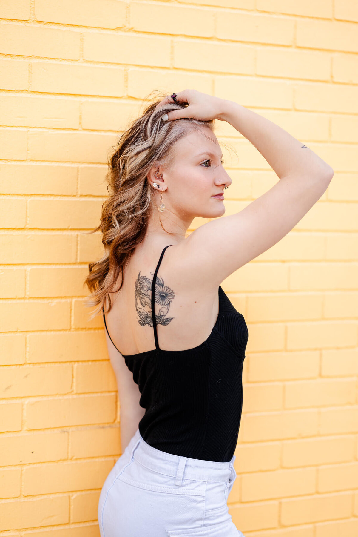 A high school senior wearing a black top and white denim skirt stands in front of a yellow brick wall. She has her hand  in her hair and is facing away to show off the tattoo on her shoulder.