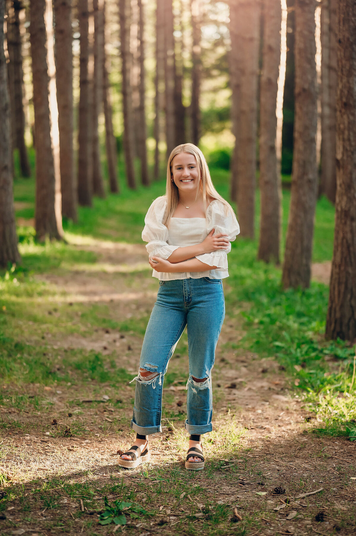 A female senior from Pewaukee High School stands amongst rows of tall trees at Delafield's Lapham Peak while wearing a peasant top and jeans.
