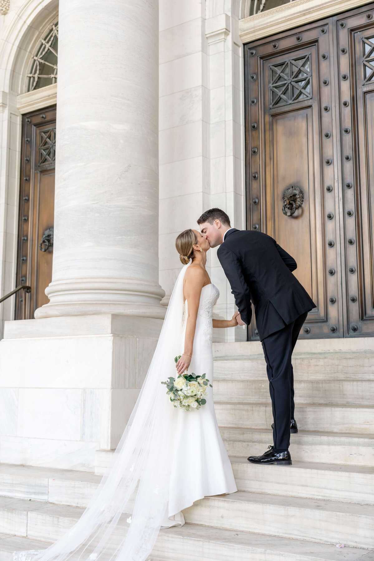 A bride and groom share a kiss on the steps of a grand building. The bride, in a white gown and veil, holds a bouquet of white flowers. The groom wears a black suit. Large doors and stone columns are in the background.