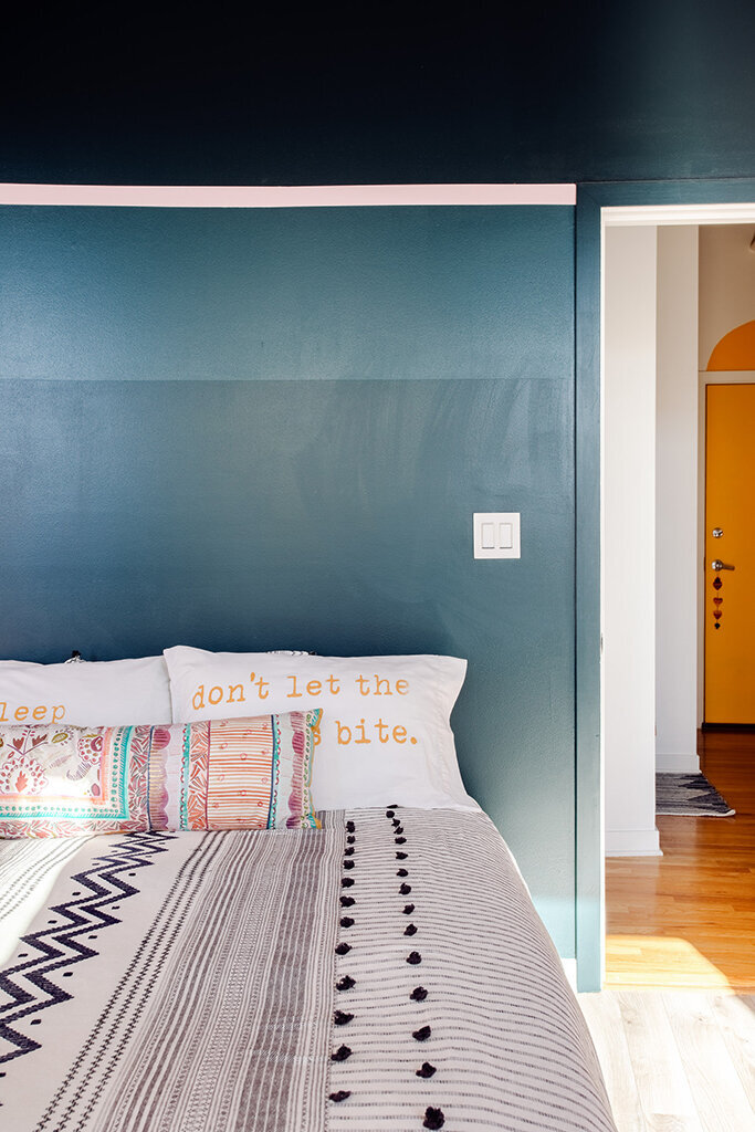 A close-up view of a bed with a patterned white and gray duvet and several pillows against a teal wall with a dark blue upper section and a thin pink stripe separating them. One white pillow has the embroidered text "don't let the bed bite." A colorful, horizontally striped lumbar pillow rests in front. A light switch is visible on the wall. A glimpse into an adjacent room shows a bright yellow arched doorway and hardwood floors.