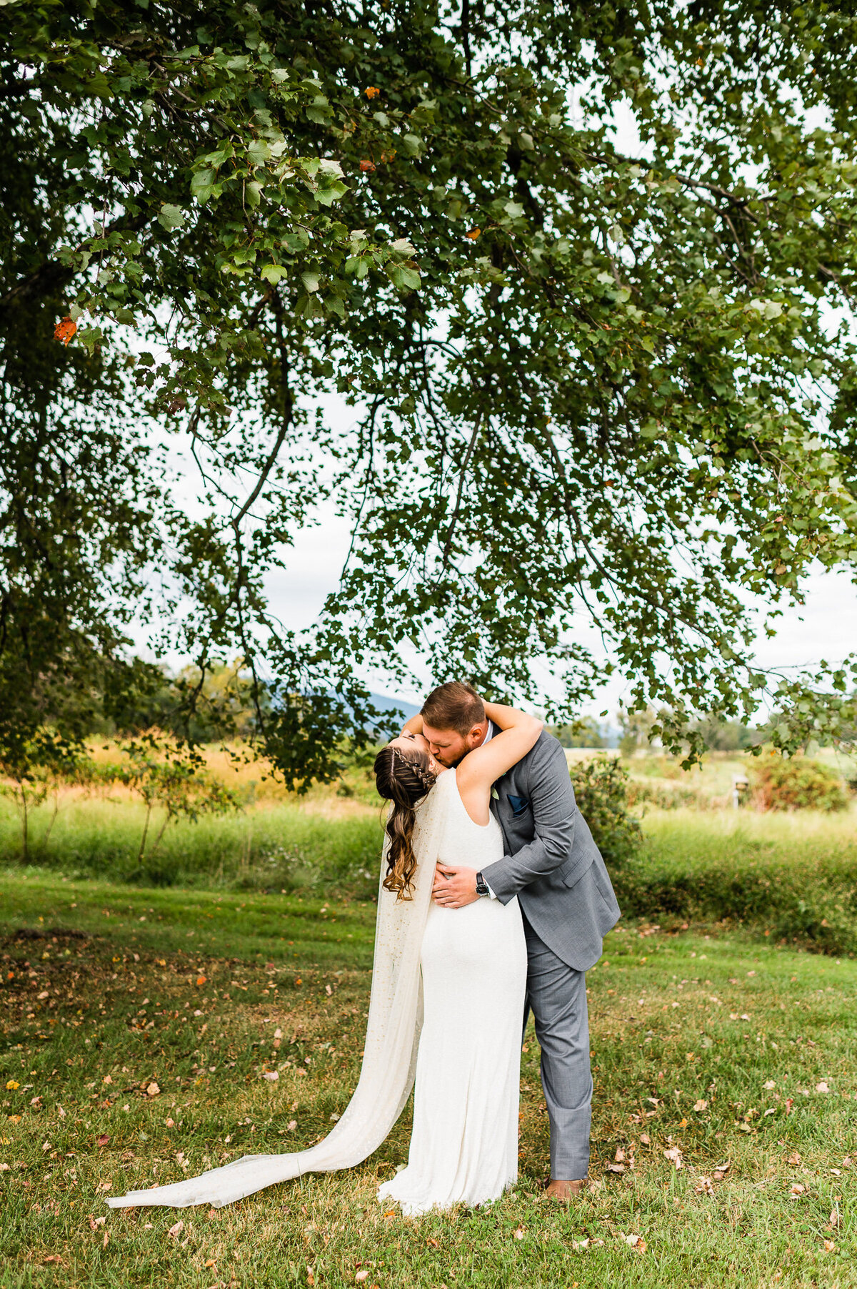bride and groom embracing on a field under trees as she groom holds the brides waist and she leans back as he kisses her neck captured by Virginia wedding photographer