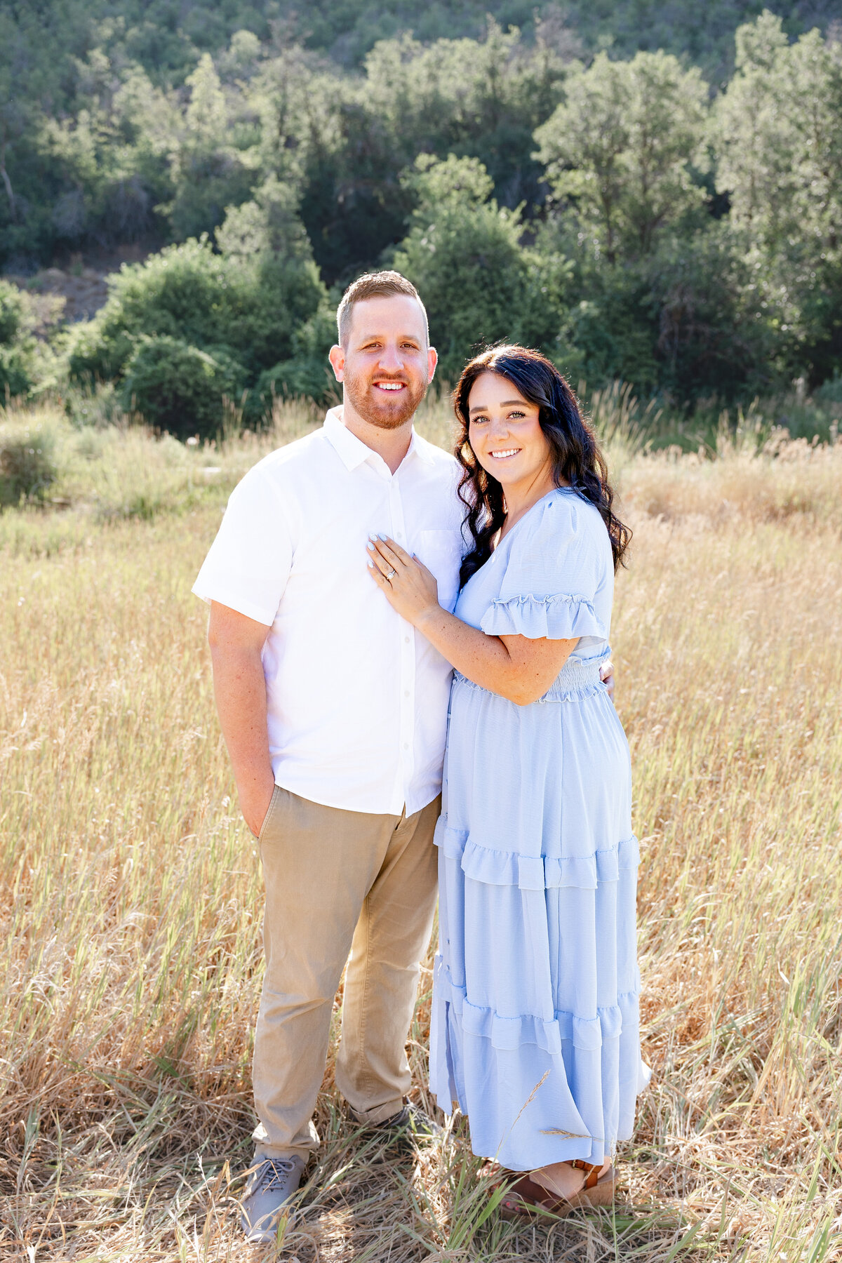 A couple stands together in a sunlit field, the woman’s hand resting on her partner’s chest. Their soft smiles and coordinated outfits make for a timeless portrait in Midway, Utah.
