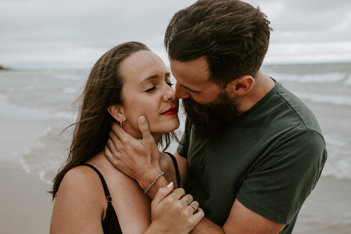 Close up image of man and woman at engagement photo shoot at Grand Bend beach. The man has his hand on the woman's neck, the woman's hand is gently holding his forearm. Their noses are touching and about to kiss.