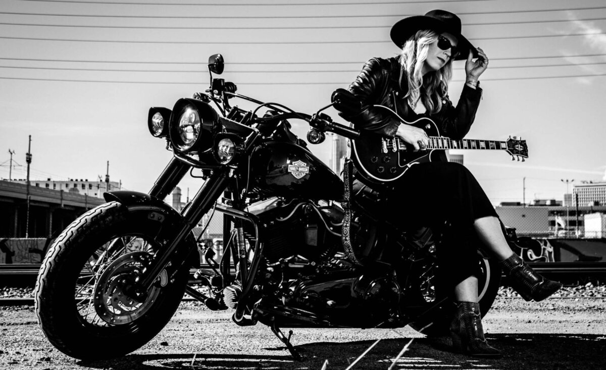 Female musician portrait Mandy Rowden black and white wearing black dress leather jacket black cowboy hat leaning against motorcycle holding guitar city bridge background