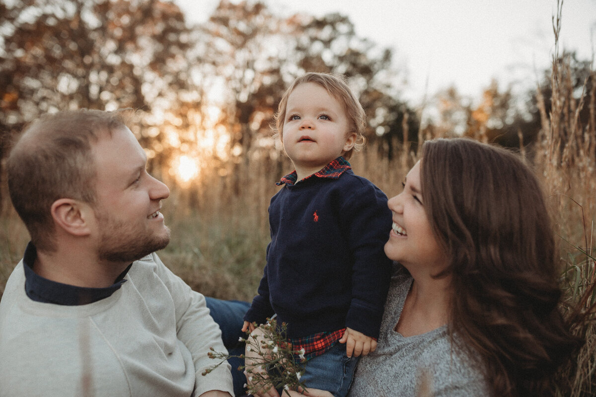 A mom and dad smiling at their cute toddler boy as the sun sets behind them. 