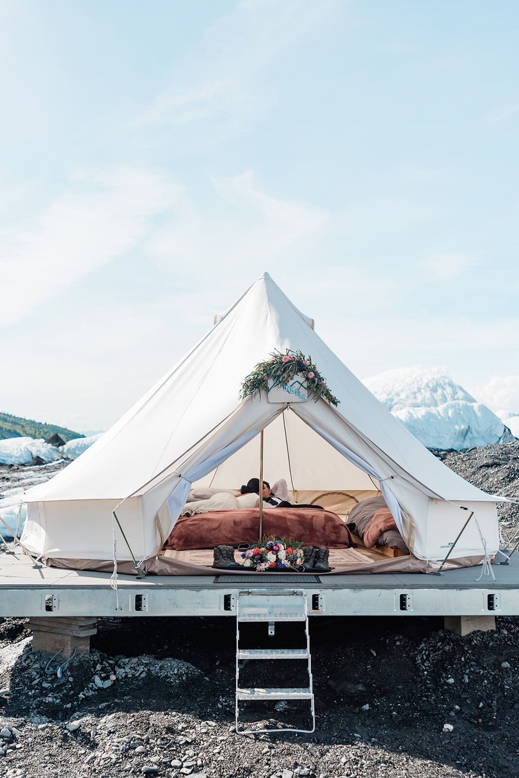 a light colored canvas bell tent on the Matanuska Glacier in Alaska is decorated with wedding flowers over the arch of the doorway during a private luxury helicopter glacier elopement. Inside you can see the comfortable luxury bedding.  Behind it in the distance rises mounds of ice and a blue and the bride and groom can just barely be seen inside the tent area as they relax in the luxury glamping space. 