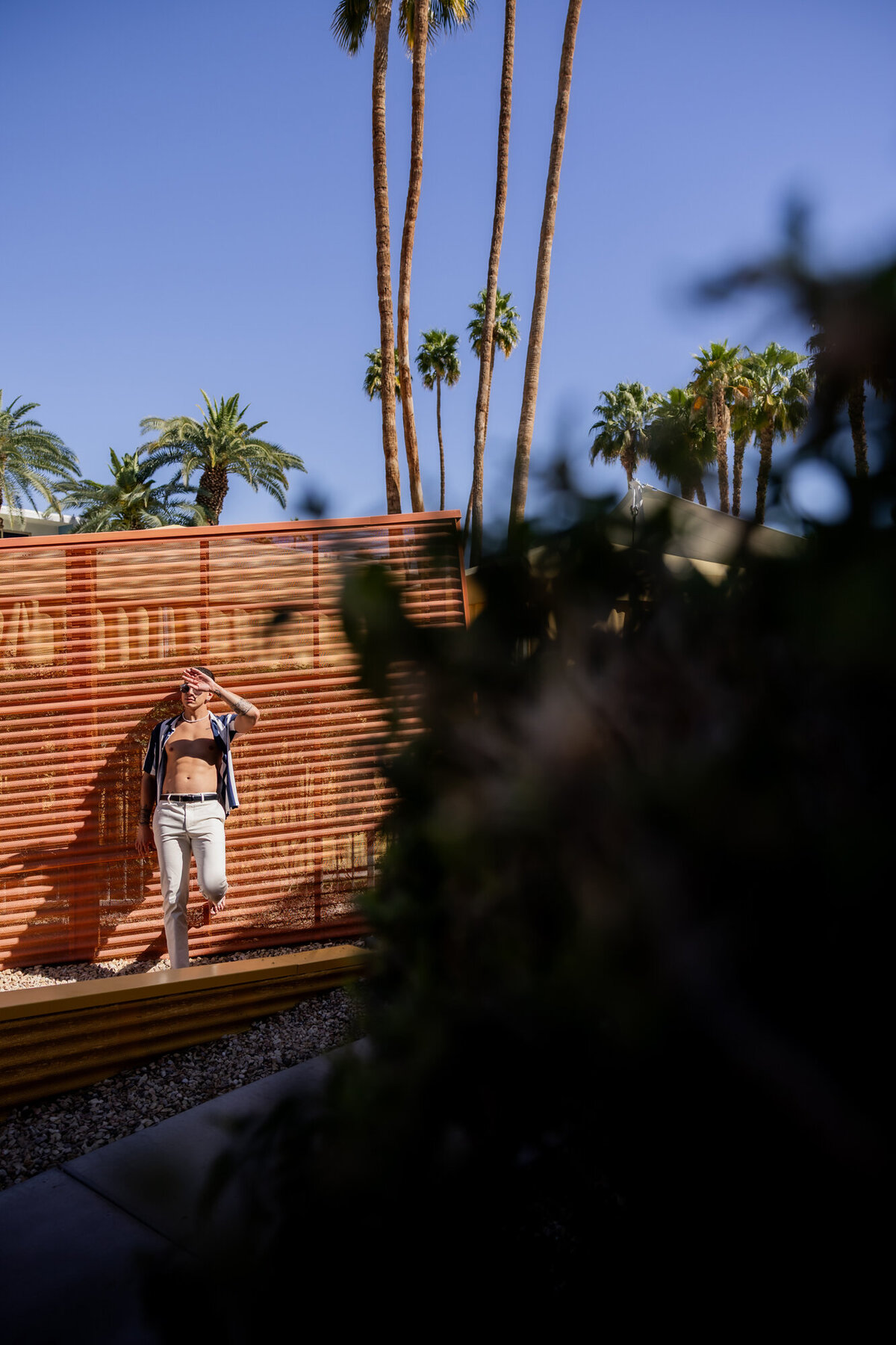 Model walking in a white outfit against a bold orange wall and desert shadows, styled editorial with Palm Springs flair.