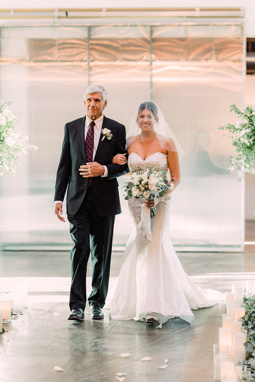 The bride, in a strapless lace wedding gown and veil, walks arm-in-arm with her father down the aisle adorned with candles and greenery, both looking forward with calm expressions.