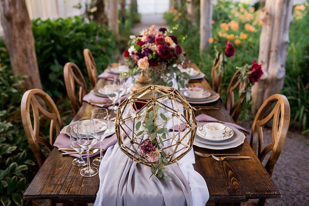 A dark wood farm table underneath a rustic wooden arbor with wood chairs that have geometric shaped backs sit in the gardens at Carnton Plantation.  The farm table has a soft gray organza table runner with large gold geometric spheres. A large floral centerpiece in a gold footed bowl had king protea, burgundy dahlia, peachtea roses, burgundy ranunculus, peach carnations and silver dollar eucalyptus. The table is set with gold flatware and stacked plates. The plates are a scalloped edge charger, a gray dinner plate, a white salad plate with gold geometric patterns topped with a white bowl holding a prince protea with a purple napkin.