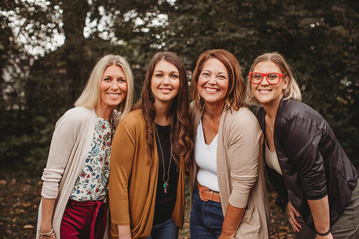 Four female mental health professionals from a therapy center's leadership team stand together outdoors, smiling warmly at the camera. They're wearing casual professional attire in earth tones and autumn colors, including cardigans and layered clothing, against a natural wooded background with bokeh lighting effects.