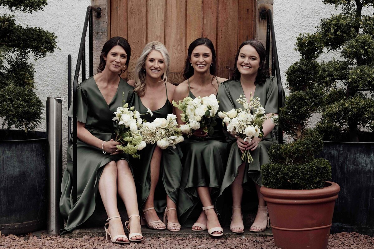 A group of four bridesmaids sit on a step at Prestonfield House. Each holds a bouquet of flowers