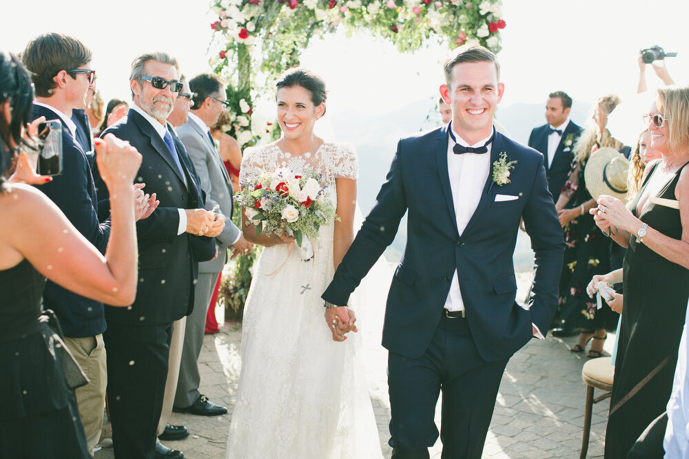 A newlywed couple walks hand-in-hand down the aisle, beaming with joy as guests celebrate their beautiful outdoor wedding with a stunning mountain backdrop.