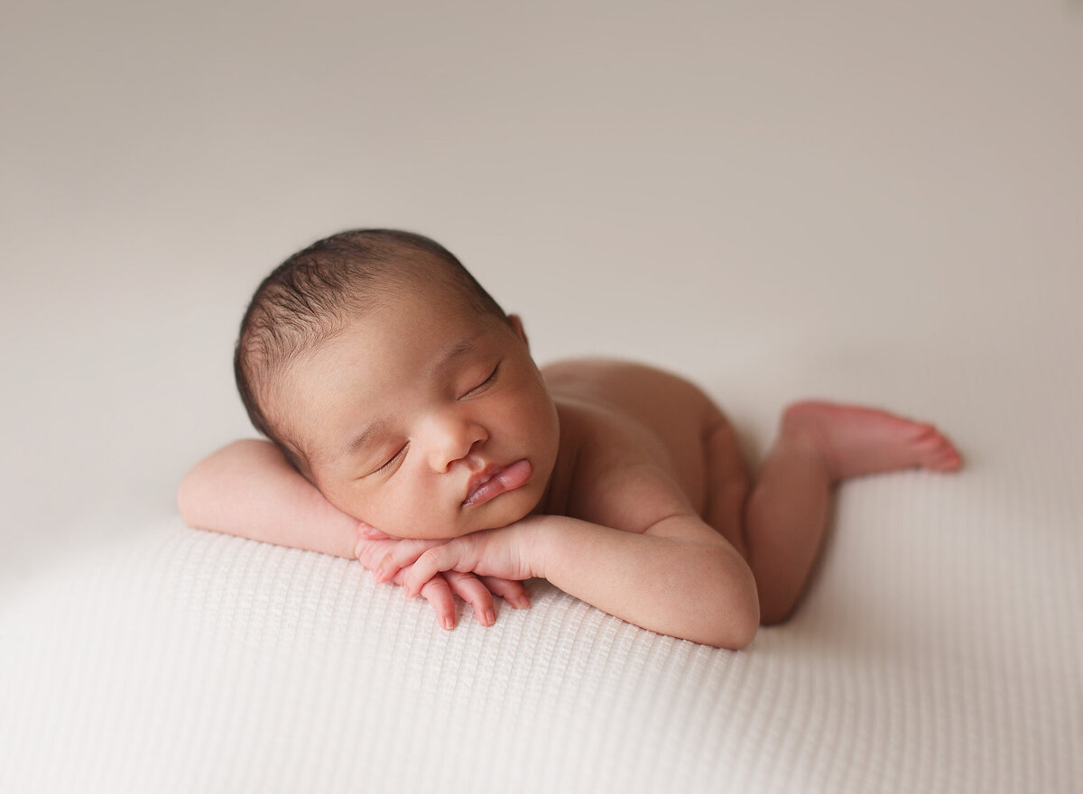 Newborn boy is posed on a white textured backdrop, posed on his tummy and facing forward towards the camera with his hands under his chin and his left foot kicked out in the back