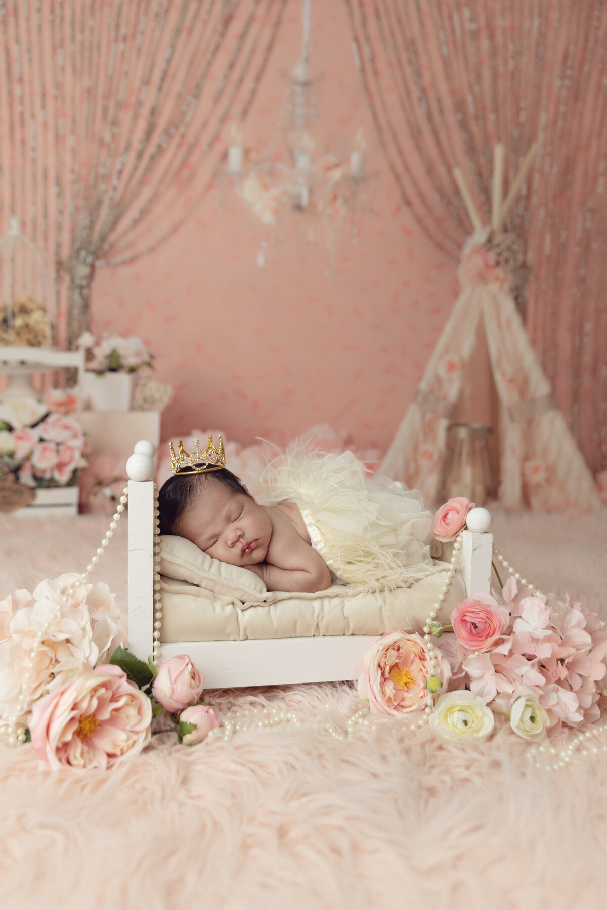 A newborn baby girl princess sleeps in a tiny white bed surrounded by flowers wearing a golden crown
