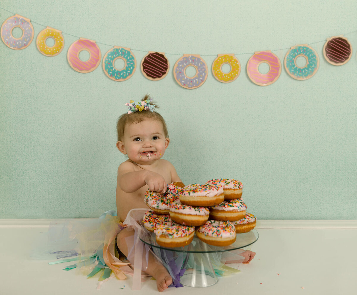 1 year old girl sitting with a pile of donuts for her birthday. Donut banner hanging behind her.