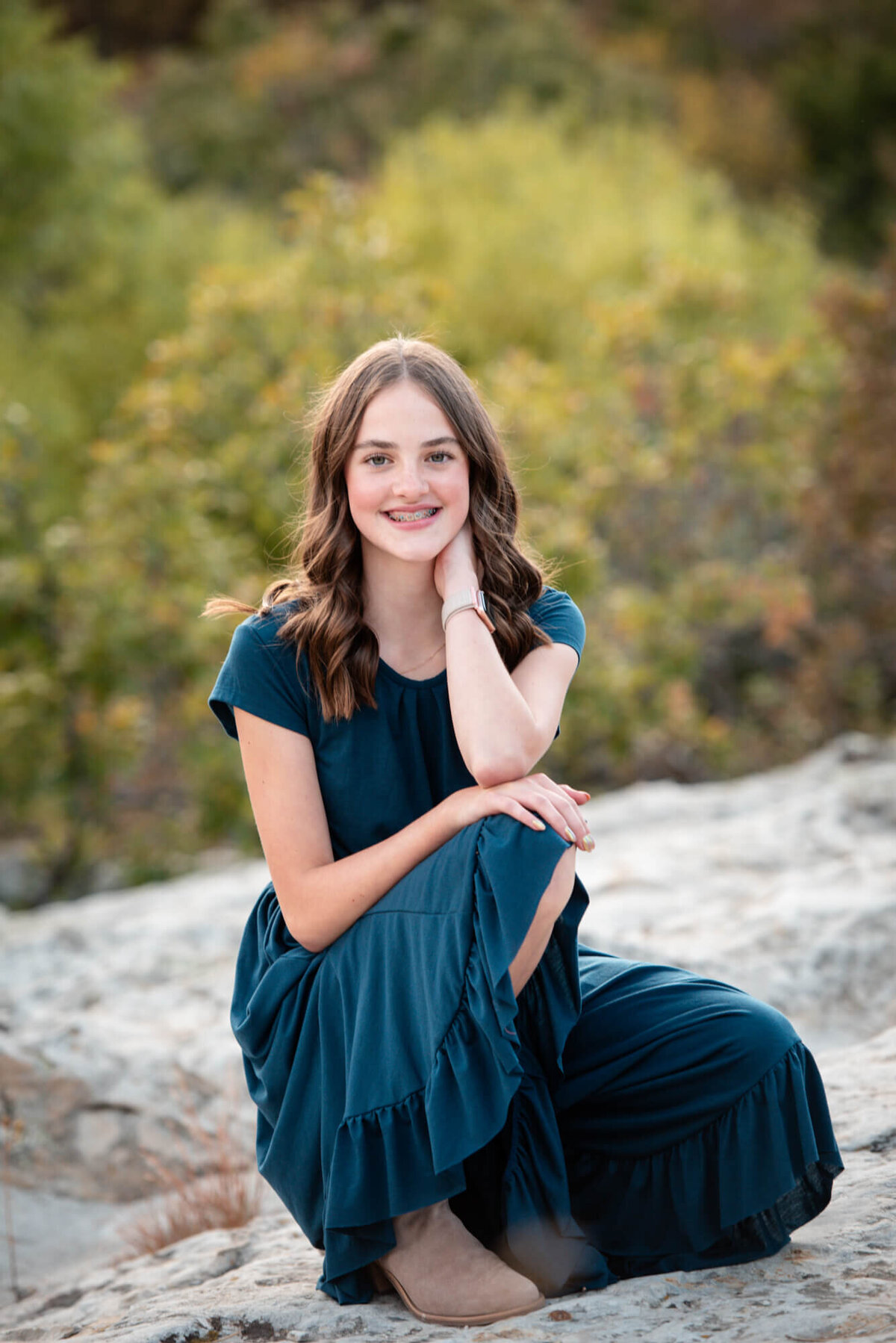 A teenage girl in a blue dress kneels on a large rock at sunset while smiling