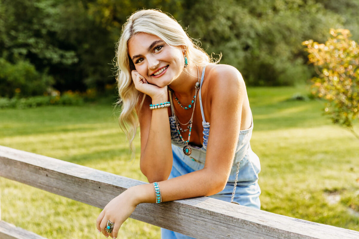 high school girl in a denim jumpsuit and turquoise jewelry leaning forward on a fence while resting her chin on one hand.
