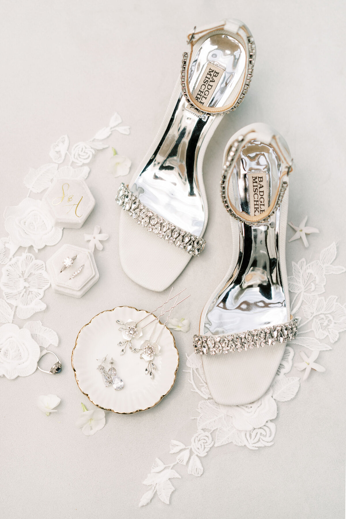 An overhead view of bridal accessories, perfect for a Banff Alberta wedding, includes bejeweled open-toe heels, earrings, a ring in a hexagonal box, and a necklace on a small dish, all arranged on a lace cloth.