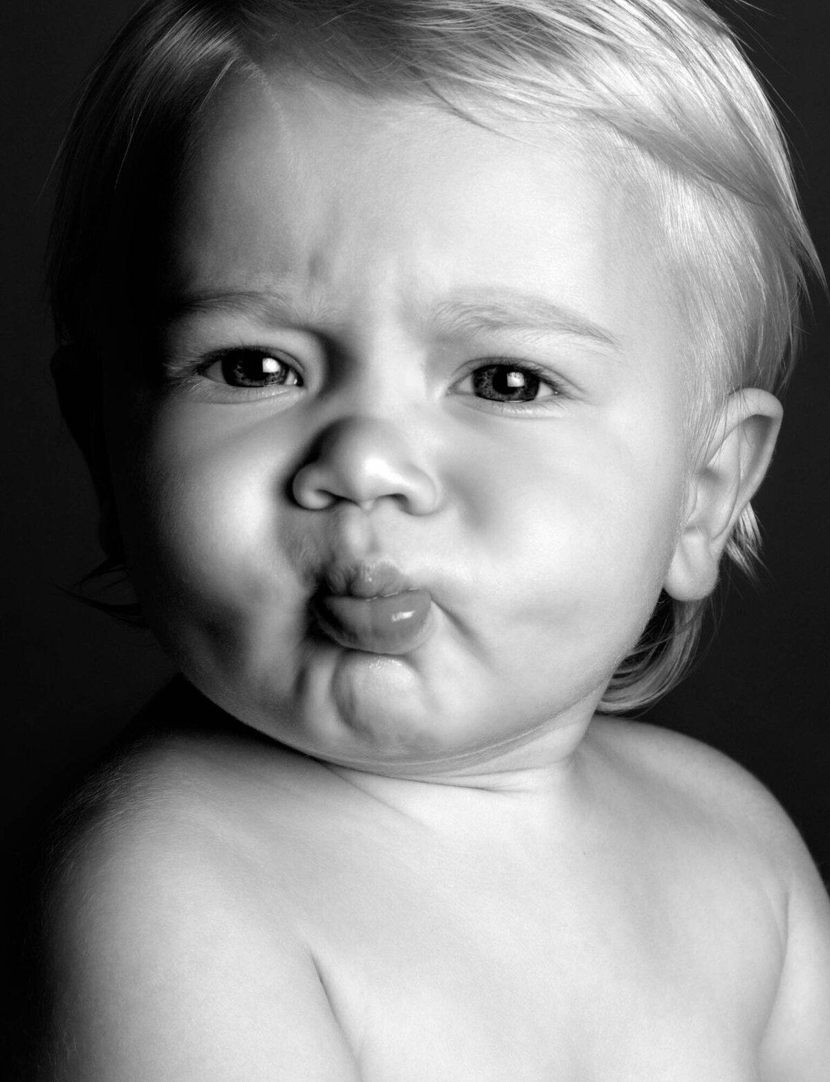 A black and white photo of a baby with light hair, making an exaggerated pouty face. The expression is playful and humorous, showcasing plump cheeks.