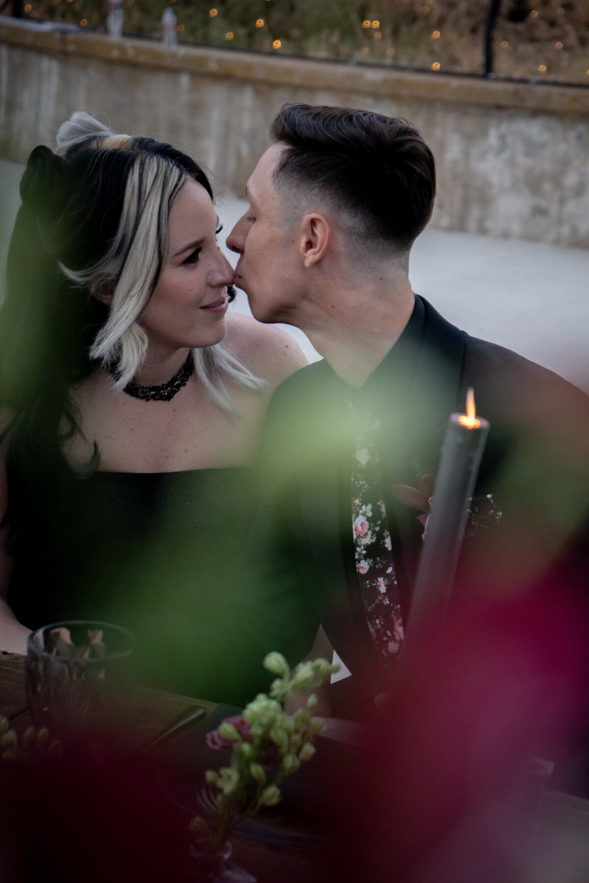 Closeup portrait of bride and groom at their mountain Oak Glen wedding table during twilight, the groom gently kissing the bride's nose in their goth attire