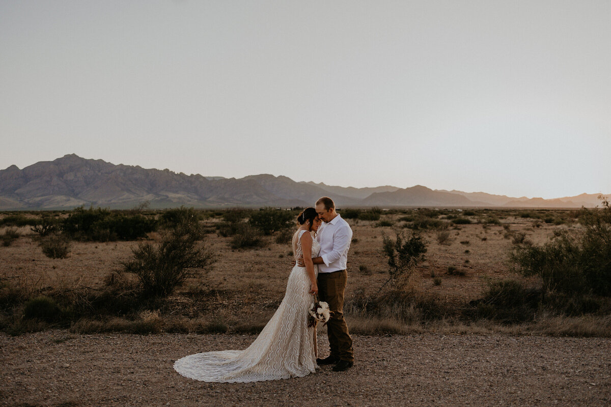 bride and groom holding each other in front of a mountain as the sun goes down