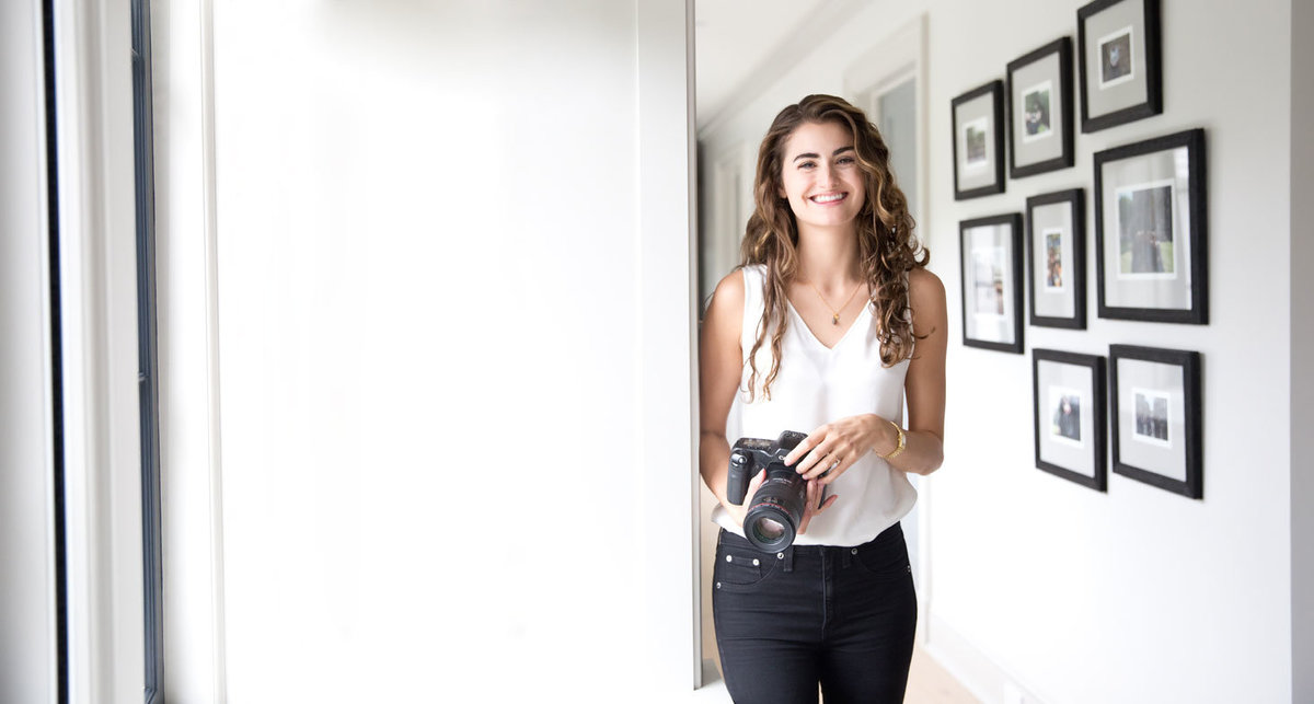 Laura Volpacchio smiles for the camera while holding her camera  in hallway with framed wall gallery in Sag Harbor, NY.