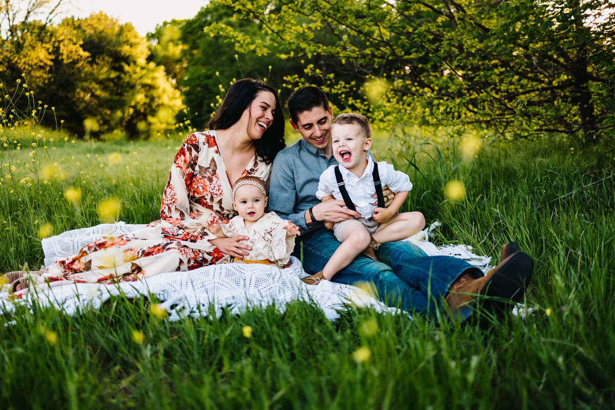 Maternity and family photography trends in Albuquerque showcase this professional capture of a family on green grass adorned with small yellow flowers. The man is holding a baby who is crying, while the woman, dressed in a white dress with orange flowers, looks on.