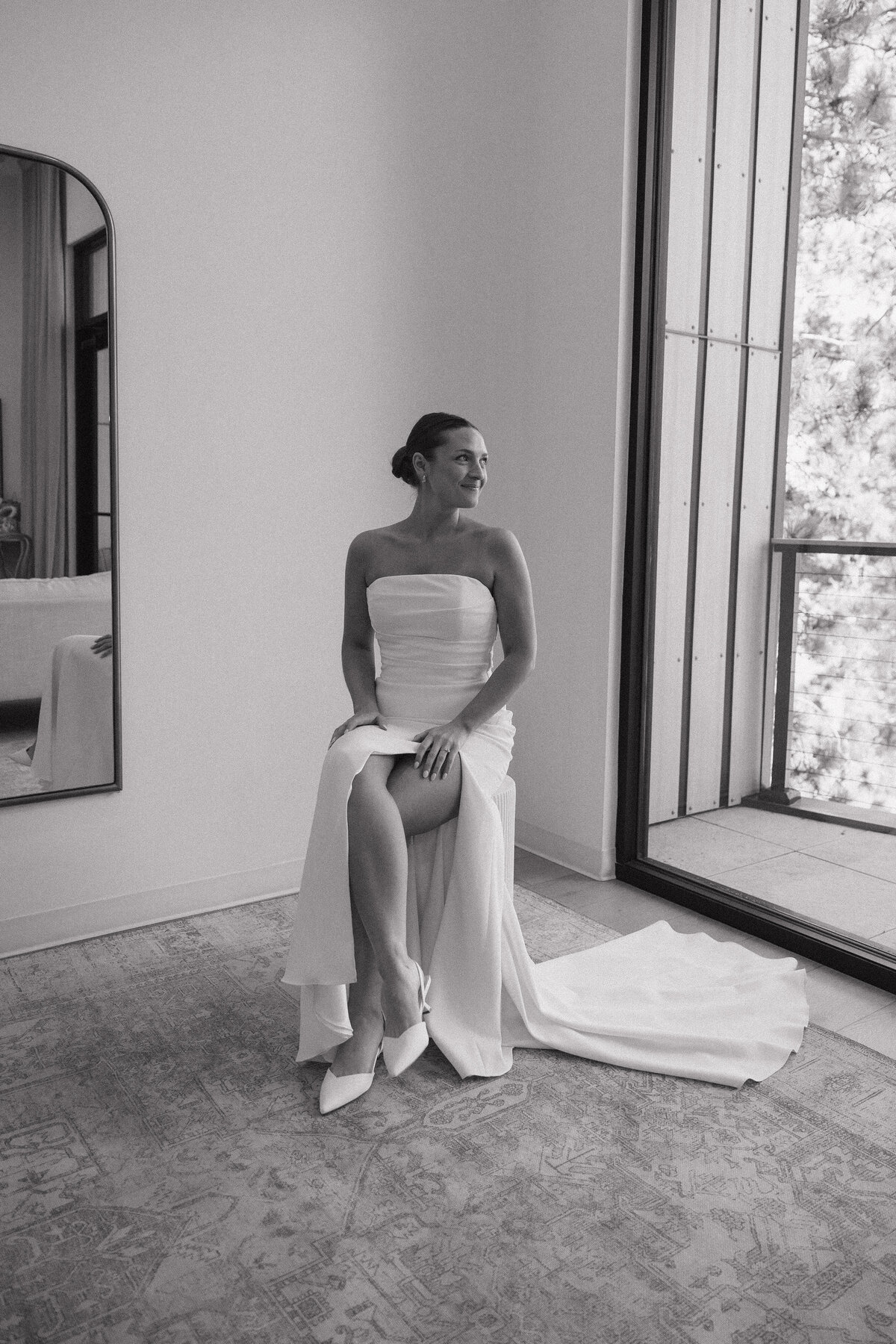 A bride in white satin strapless dress with a large leg slit sits on a cylindrical stool with both hands resting on her upper thigh. She looks over her left shoulder out a very large window that reveals a small balcony. Her face is stoic and she has a slicked back bun and small gold hoop earrings in.  