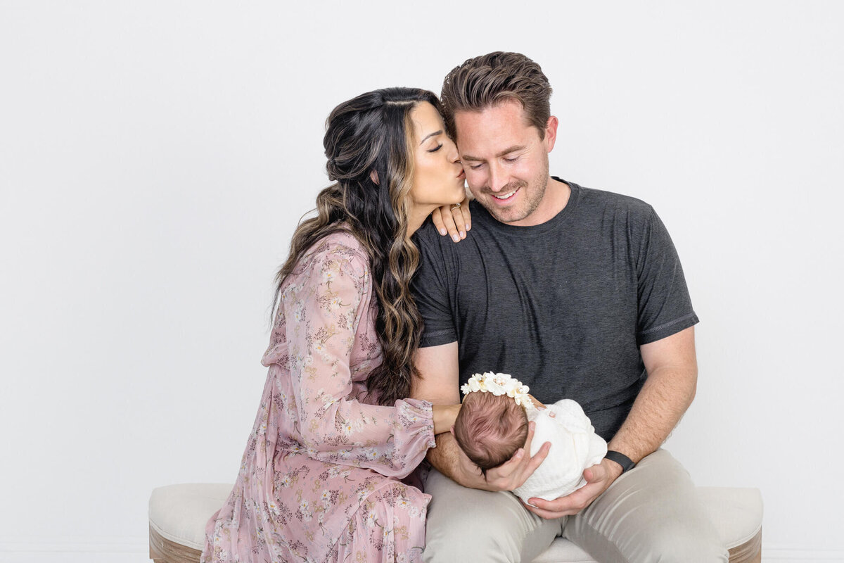 A woman in a floral dress kisses a smiling man on the cheek as he holds a newborn baby wrapped in a blanket with a flower headband. They sit together on a light-colored bench against a plain white background.