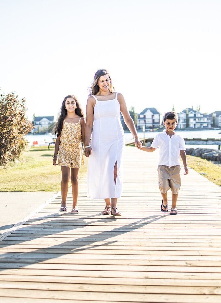 A mom in a white dress is walking near a lake holding her son and daughter's hands
