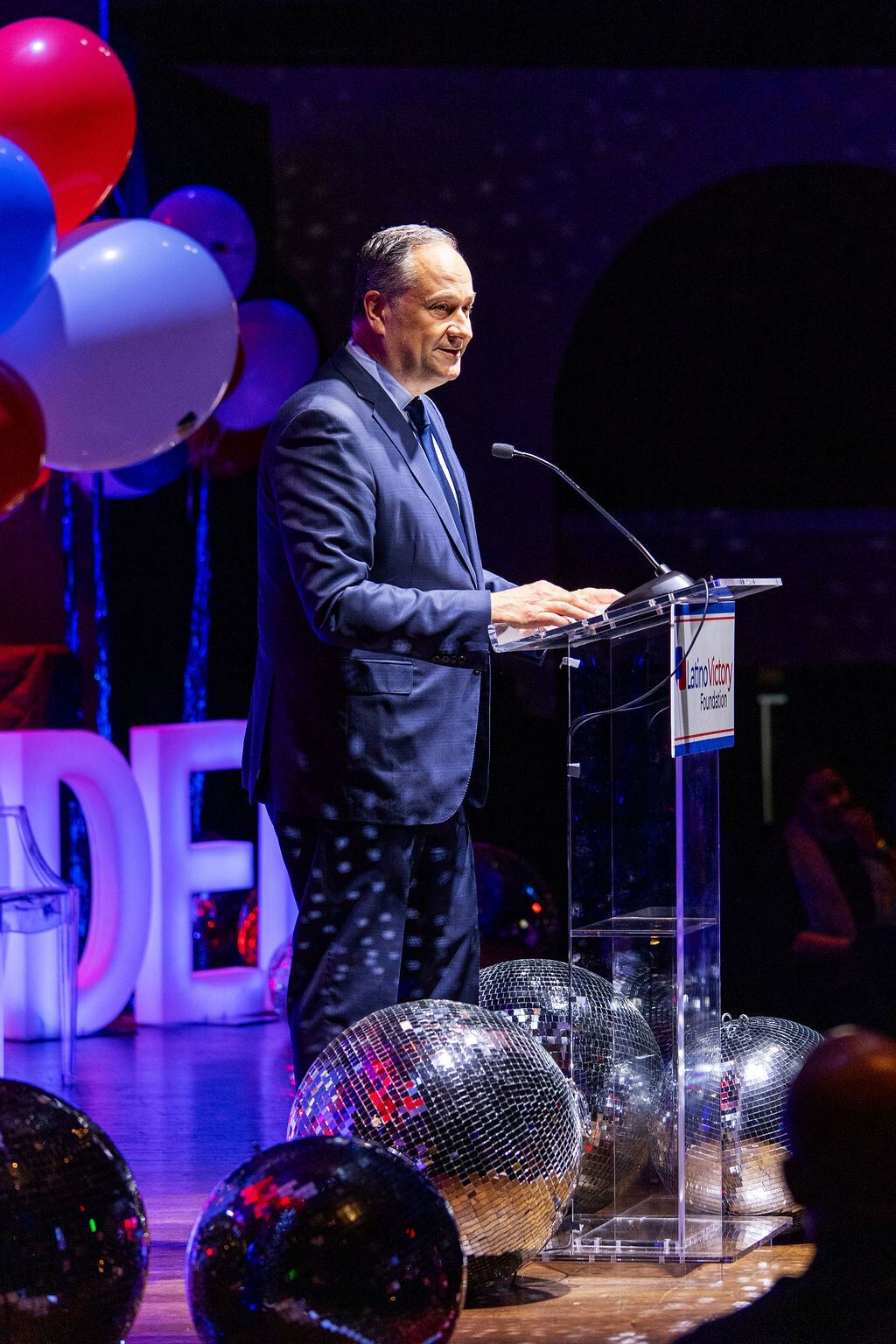 A person speaks into a microphone with red, white, and blue balloons around them.