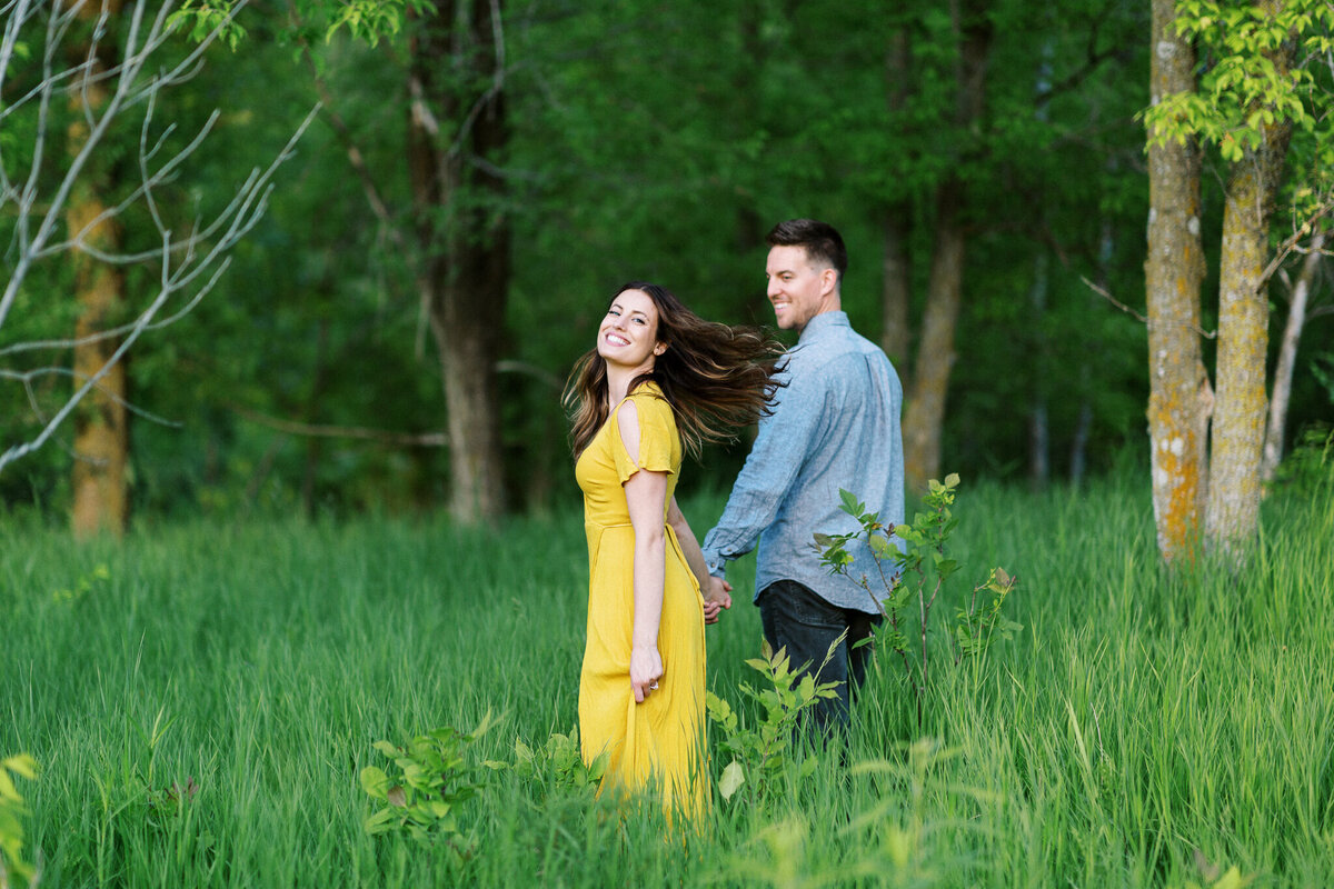 A couple walks arm in arm in Maple Grove, sharing a joyful moment  while she looks at the camera.