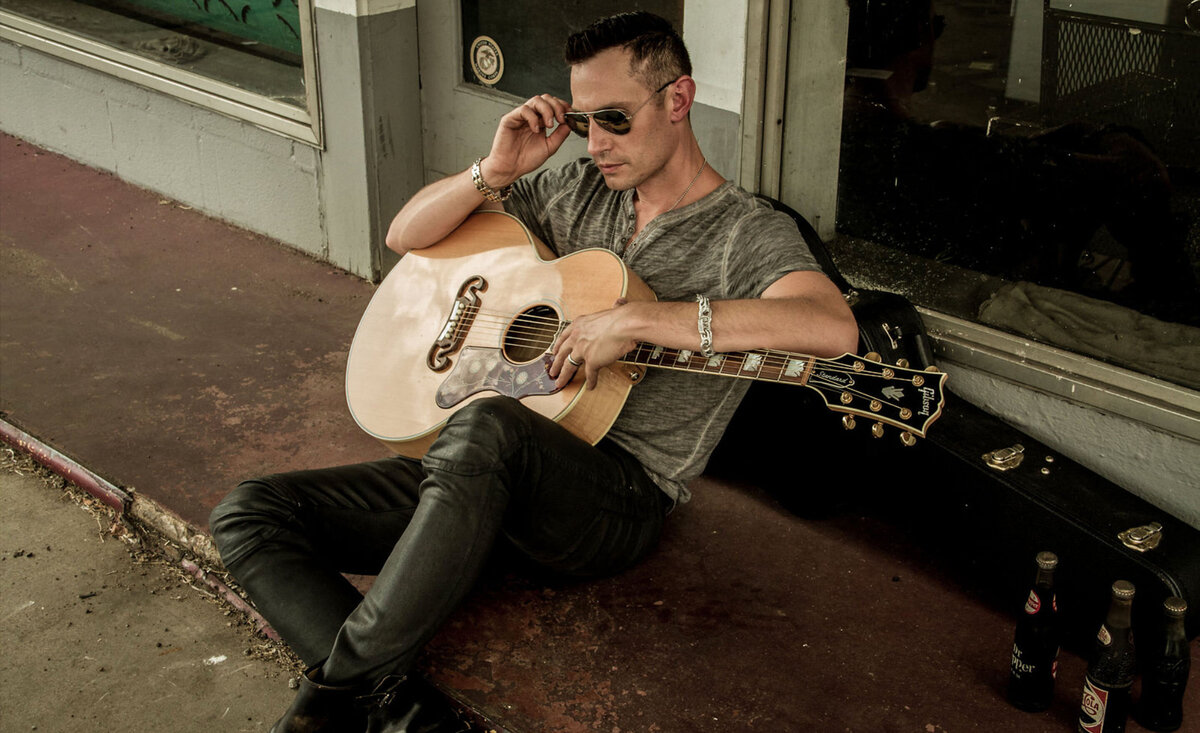 Male musician photo Adam Rutledge wearing grey t shirt black jeans sunglasses sitting against guitar case holding pale wood guitar