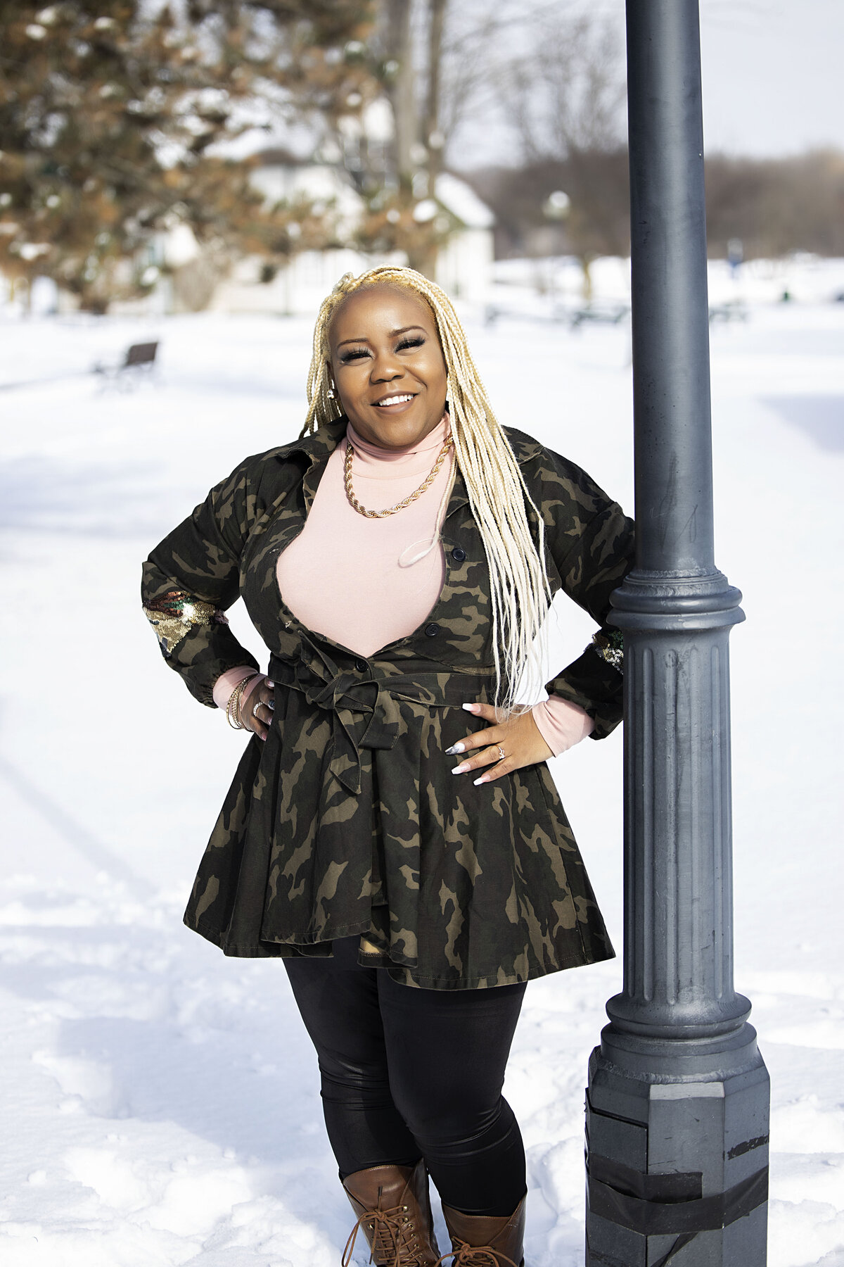 African American woman in camouflage jacket, pink turtleneck brown boots, and black jeans standing next to light pole