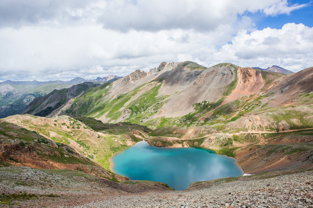 A bright blue  alpine lake sits nestled in green mountains with orange  rocky crests.