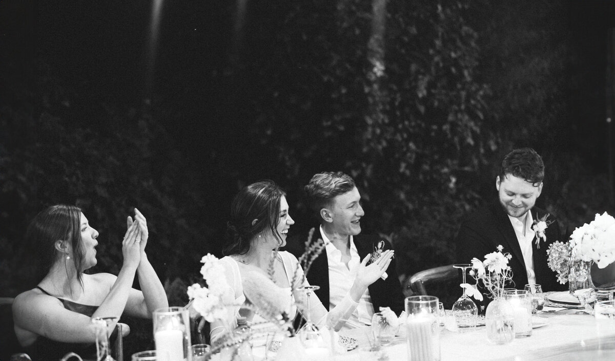 Bride and groom standing at an ivy covered alter laughing as groom puts wedding band on bride's finger