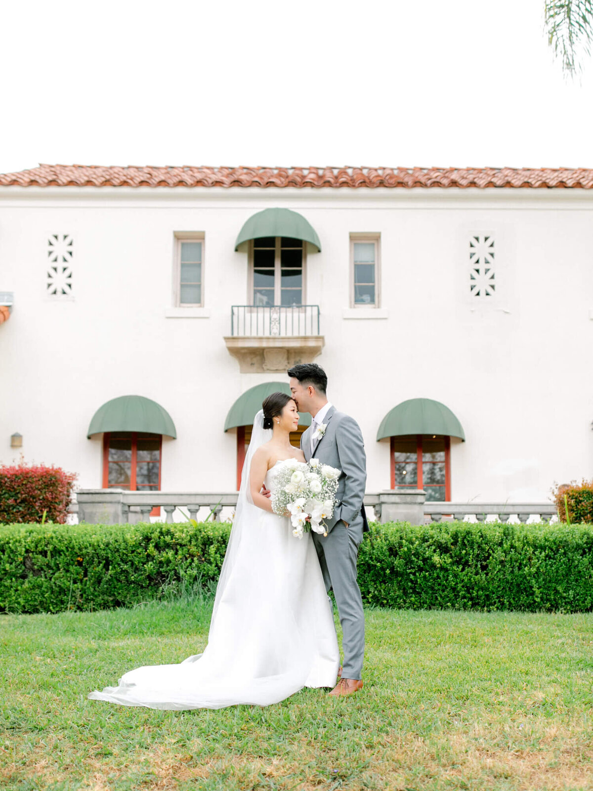 Bride and groom embrace on a lush lawn in front of a Mediterranean-style villa with arched windows. The scene is serene and romantic.